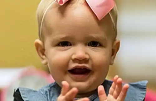 Little girl student with bow in hair at La Petite Academy clapping and smiling at camera; daycare summer camps