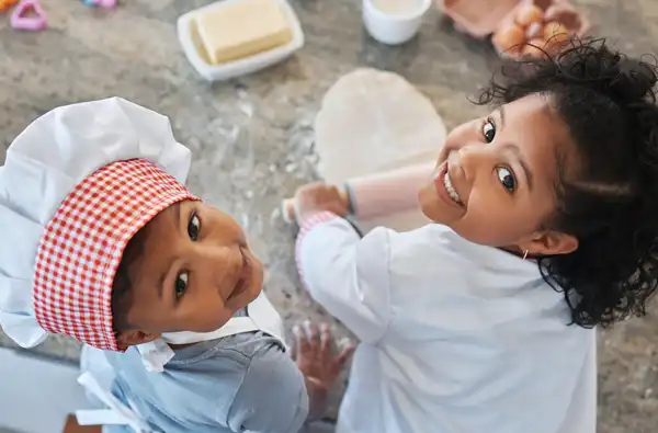 Cooking With Kids: Life Skills That Start in the Kitchen blog article main image; two kids looking up to camera while learning to cook.